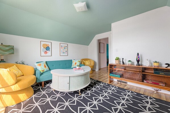 Un salon avec un canapé de couleur turquoise, un fauteuil jaune et une table basse ronde. Deux œuvres d’art encadrées ornent le mur, et une bibliothèque contient divers objets décoratifs.