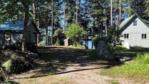 A cabin with a metal roof, surrounded by tall trees and a gravel path. - Cozy, Off-Grid Camp on Spectacle Pond w/ private water frontage! (Osborn)