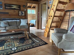 A cozy cabin interior with a sofa, a coffee table, a rug with animal prints, and a wooden staircase. - Cozy, Off-Grid Camp on Spectacle Pond w/ private water frontage! (Osborn)