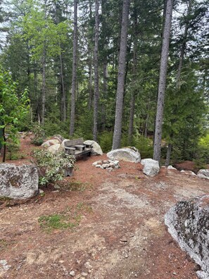 A forested area with a wooden bench and large rocks. - Cozy, Off-Grid Camp on Spectacle Pond w/ private water frontage! (Osborn)