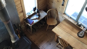 A cabin interior with a wood stove, a dining table with chairs, a window, and a small table with items on it. - Cozy, Off-Grid Camp on Spectacle Pond w/ private water frontage! (Osborn)