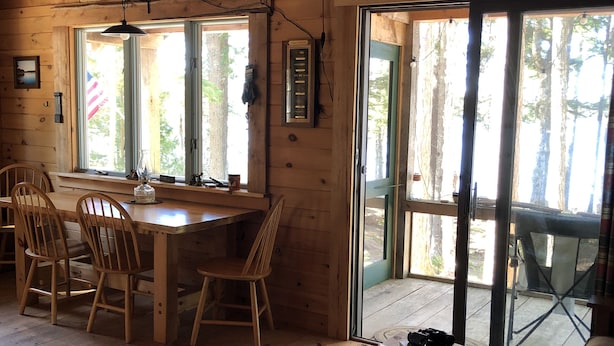 A wooden cabin interior with a dining area, a sofa, and a large window. - Cozy, Off-Grid Camp on Spectacle Pond w/ private water frontage! (Osborn)