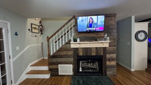 A living room with a wooden staircase, a fireplace, a mounted television, and a clock on the wall. - Bright Aqua Yacht Harbor Pickwick Lake Lakeview Home Boat Garage Sleeps 10+ (Iuka)