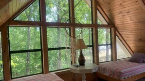 A room with two beds, a small table with a lamp, and large windows offering a view of trees. - Bright Aqua Yacht Harbor Pickwick Lake Lakeview Home Boat Garage Sleeps 10+ (Iuka)