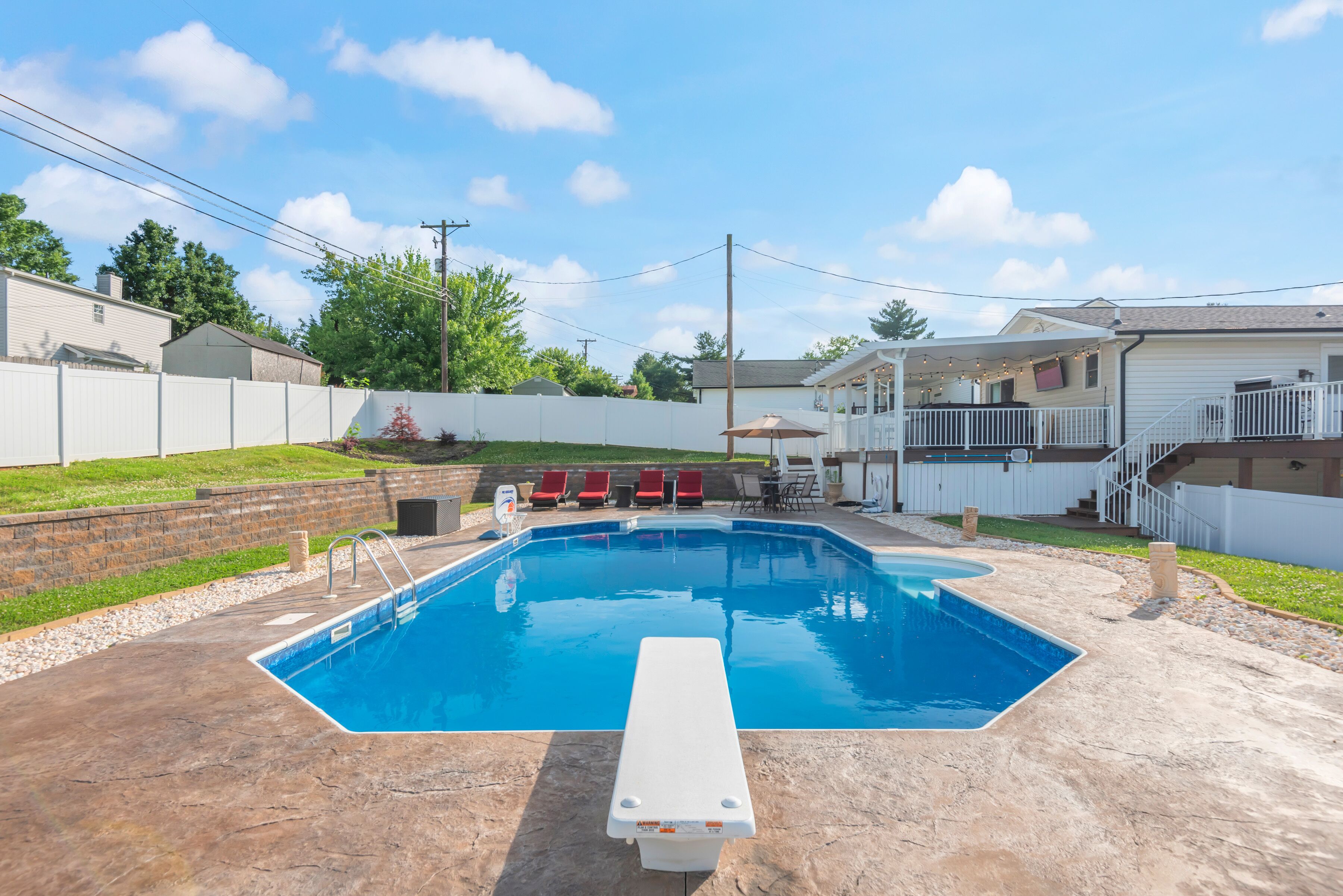Piscina residencial con tabla de salto, rodeada por una cerca blanca y un área de patio techada con luces colgantes.