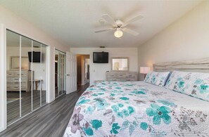 Ein Schlafzimmer mit einem großen Bett, einem Deckenventilator, einem an der Wand befestigten Fernseher und einer Schiebetür zu einem weiteren Raum.