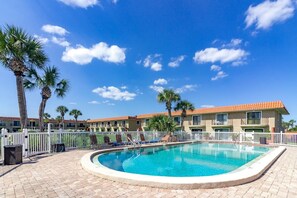 A swimming pool with lounge chairs, palm trees, and a row of buildings with red-tiled roofs.