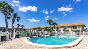 A swimming pool with lounge chairs and palm trees, surrounded by a white fence and a building with a red roof. - Seabreeze Escape at Ponce Landing 34 (St. Augustine)