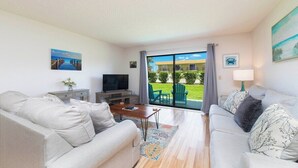 A living room with two sofas, a TV, and a coffee table. There is a large window with a view of a building and greenery. - Seabreeze Escape at Ponce Landing 34 (St. Augustine)