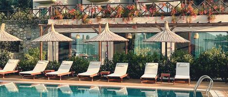 A poolside area with lounge chairs and umbrellas, a large pool, and a building with multiple windows and balconies.