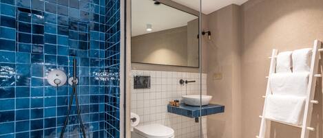 A bathroom with a blue-tiled shower, a white toilet, a sink with a mirror above it, and a towel rack with towels.