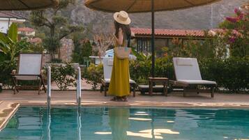 A pool area with lounge chairs and umbrellas, a woman in a yellow dress, and mountains in the background.