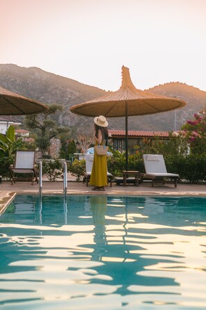 A pool area with lounge chairs and umbrellas, a woman in a yellow dress, and mountains in the background. - Boho Garden Selimiye (Marmaris)