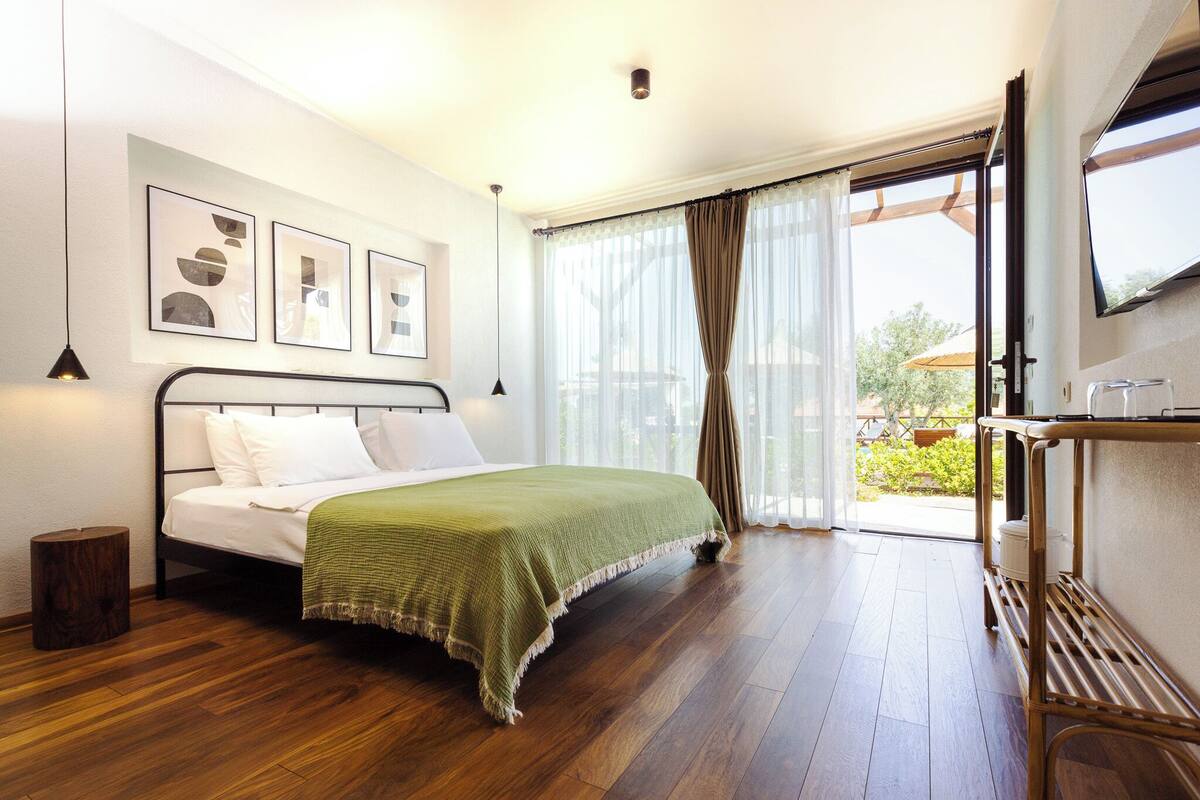 Een slaapkamer met een bed, een houten vloer, een televisie aan de muur en een raam met gordijnen.
