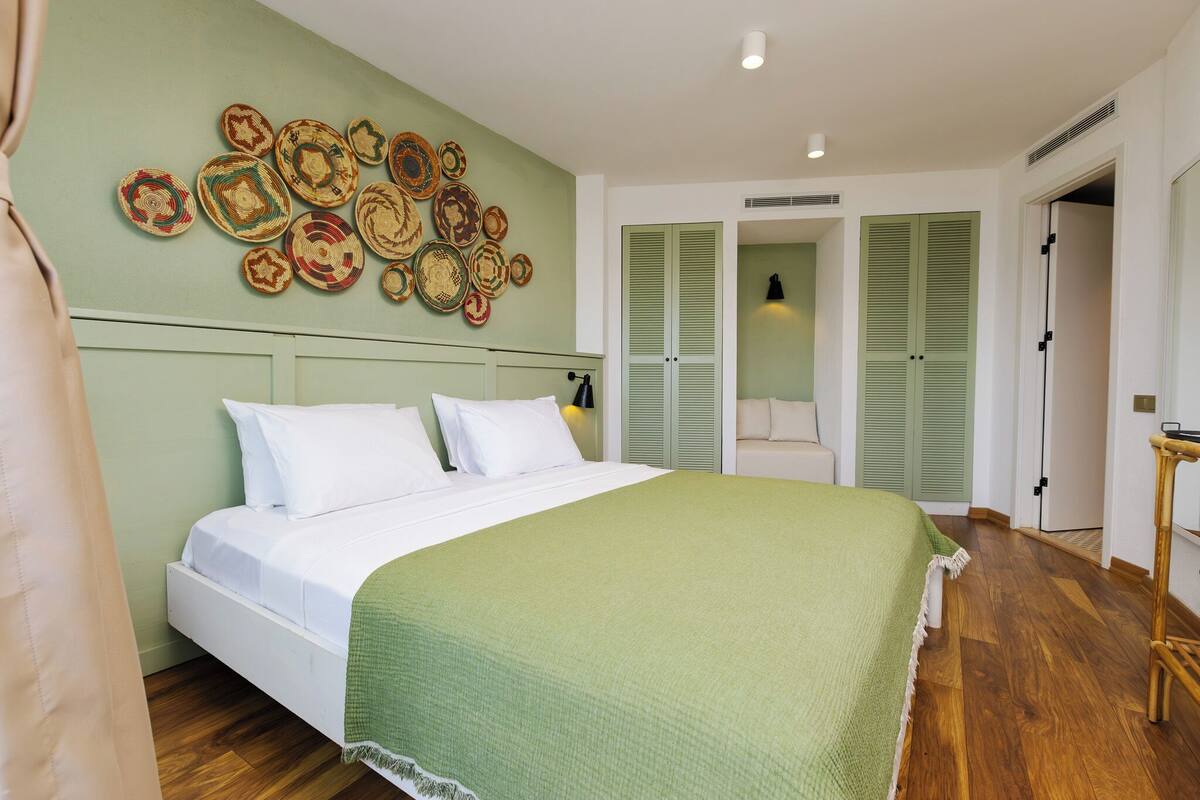 Een net opgemaakt bed met een groene deken, een wand met geweven kunst en een houten vloer.
