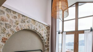 A bedroom with a stone wall, a bed with white linens, a green throw blanket, a wicker chair, and a view of the sea through large windows. - Boho Garden Selimiye (Marmaris)