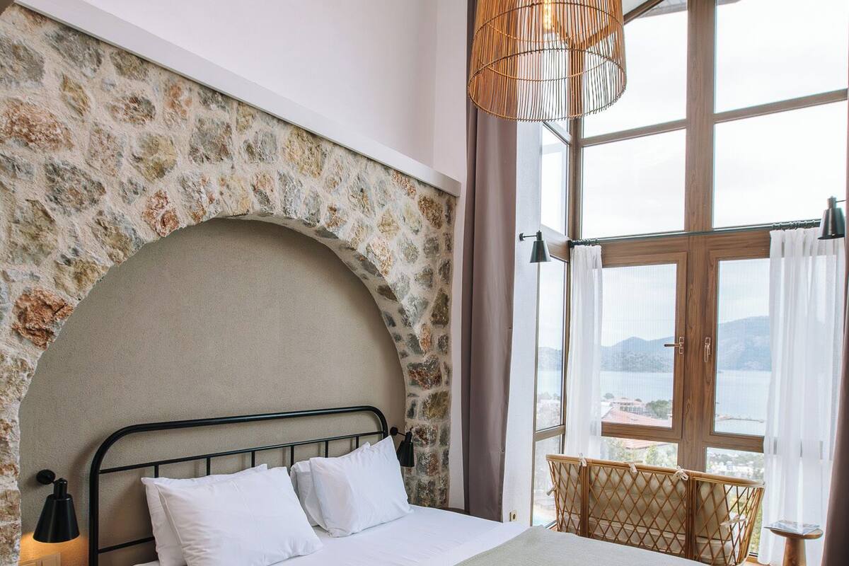 Een slaapkamer met een stenen muur, een bed met witte lakens, een groene plaids, een rieten stoel en uitzicht op de zee door grote ramen.