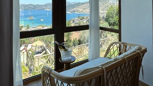 A room with a large window overlooking a scenic landscape, a wicker chair, and a table with a bottle and glass. - Boho Garden Selimiye (Marmaris)
