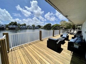 Une terrasse en bois, meublée pour l’extérieur, avec une balustrade blanche et une vue sur un lac, des bâtiments se dessinant à l’arrière-plan.