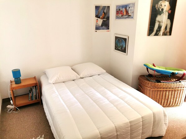 Una cama pequeña y bien hecha con edredón blanco, una mesita de madera con lámpara y una cesta de mimbre con un cochecito de bebé.
