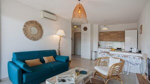 A modern living room with a teal sofa, a glass coffee table, and a wicker chair. A kitchen with white cabinets and a dining area with white chairs is visible in the background. - Playa Marina (Bastia)