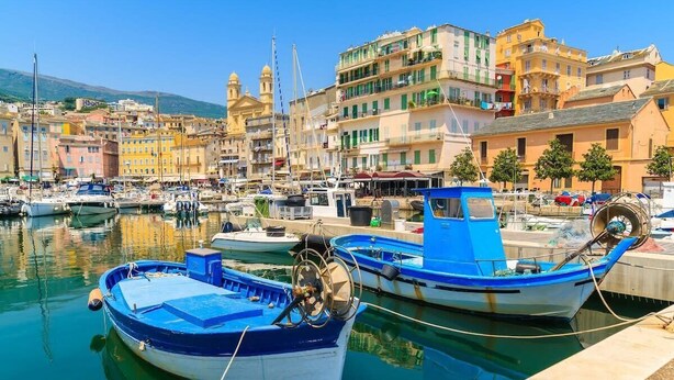 A marina with boats docked, a waterfront promenade, and colorful buildings in the background. - Playa Marina (Bastia)
