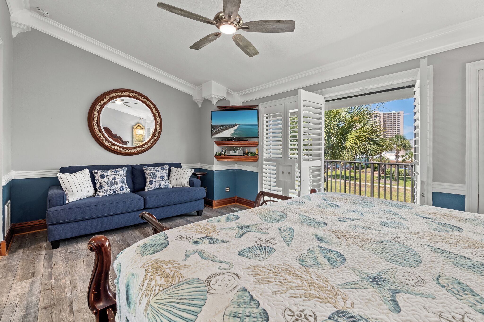 A bedroom with a bed, a blue sofa, a ceiling fan, a large window with a view of palm trees and buildings, and a wall-mounted TV.