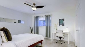 Un dormitorio con cama, escritorio, silla, ventilador de techo y ventana con cortinas.