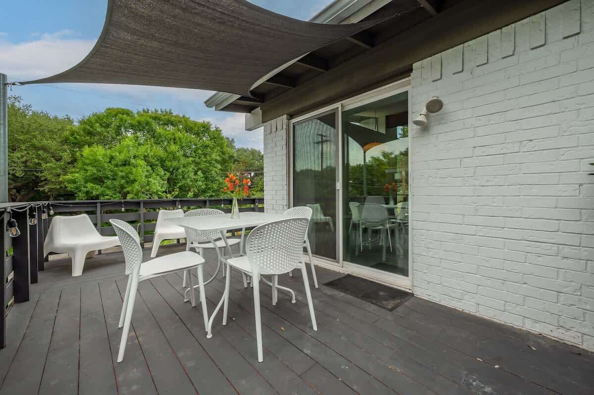Une terrasse extérieure avec des chaises et une table en plastique blanc, un auvent foncé et un mur en briques blanches agrémenté d’une porte-fenêtre coulissante.