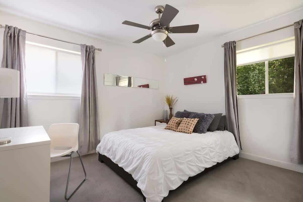 Een slaapkamer met een bed, een bureau, een stoel, een plafondventilator en een raam met gordijnen.