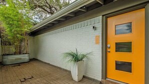 Un bâtiment à l’extérieur en briques blanches, avec une porte jaune dotée d’une fenêtre et un pot de fleurs placé devant.