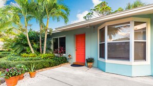 A house with a red door, a small porch, and a garden with various plants. - Coastal Cottage / 1 Mile to Hobe Sound Public Beach (Hobe Sound)