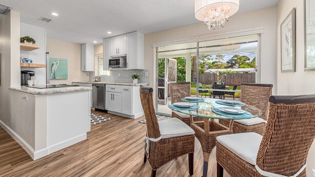 A modern kitchen with white cabinetry, a dining area with wicker chairs and a glass table, and a large sliding glass door leading to an outdoor patio. - Coastal Cottage / 1 Mile to Hobe Sound Public Beach (Hobe Sound)