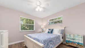 A bedroom with a bed, a ceiling fan, a nightstand, a lamp, and a cabinet with a beach scene. - Coastal Cottage / 1 Mile to Hobe Sound Public Beach (Hobe Sound)