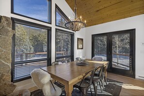 A dining area with a wooden table, chairs, and a chandelier. There are large windows with blinds, a stone wall, and a view of trees.