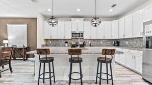 A modern kitchen with white cabinets, a granite countertop, and a stainless steel refrigerator. There are three bar stools with wooden seats and black metal frames.