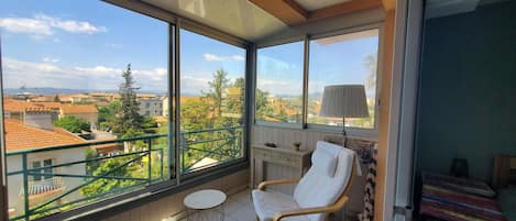 A balcony with a view of buildings and trees, a chair, a small table, and a lamp.