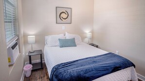 Ein ordentlich eingerichtetes Schlafzimmer mit einem Bett, zwei Nachttischlampen, einem kleinen Beistelltisch, einem Mülleimer und einer wandmontierten Klimaanlage.