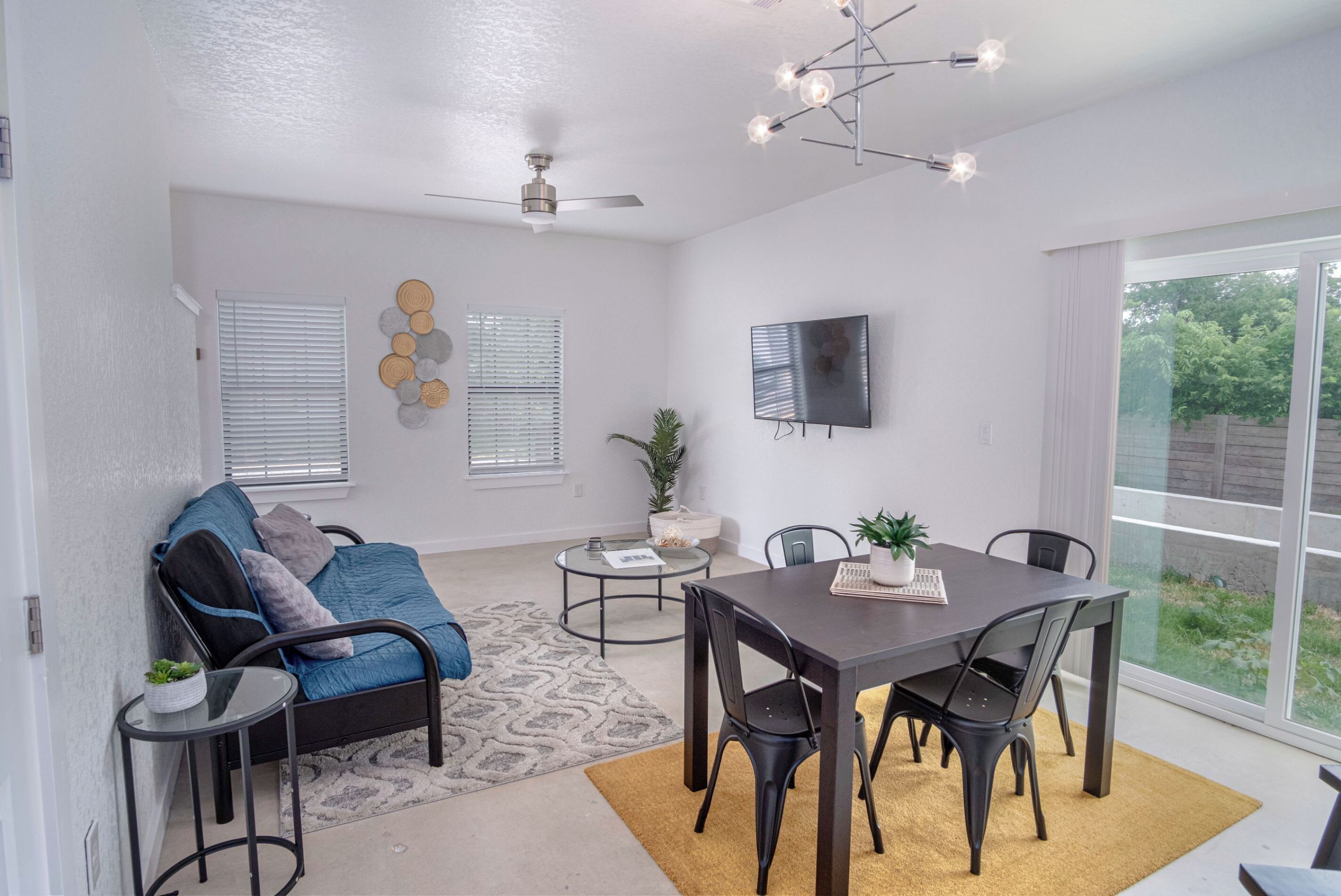 Un salon moderne avec un canapé, une table basse et une table à manger. On y trouve un ventilateur de plafond, un téléviseur fixé au mur et une grande fenêtre avec des stores.