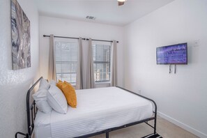 Un lit simple avec une literie blanche et un oreiller jaune, un téléviseur à écran plat fixé au mur et une fenêtre avec des stores.
