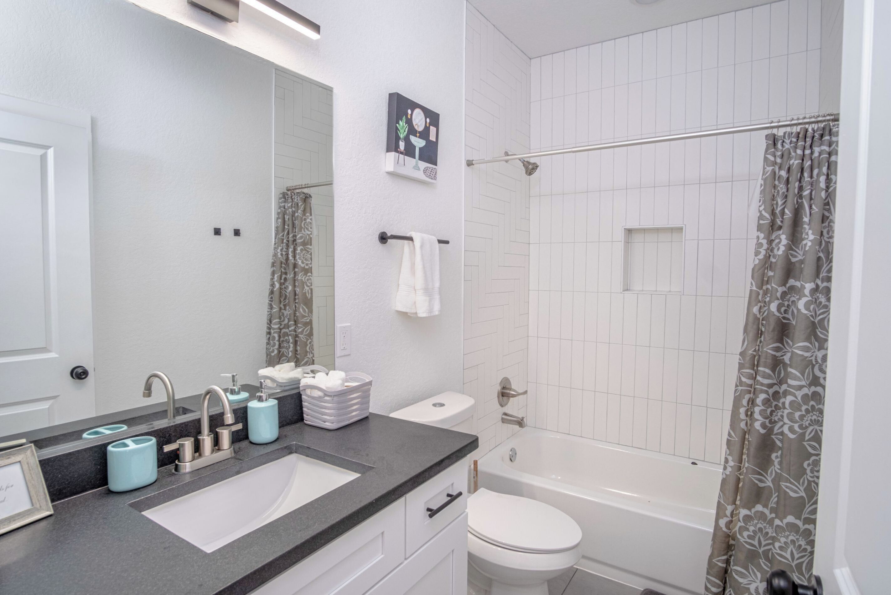Une salle de bain moderne avec un lavabo sur un plan de toilette noir, des accessoires blancs, un grand miroir et une douche agrémentée d’un rideau à motifs floraux.