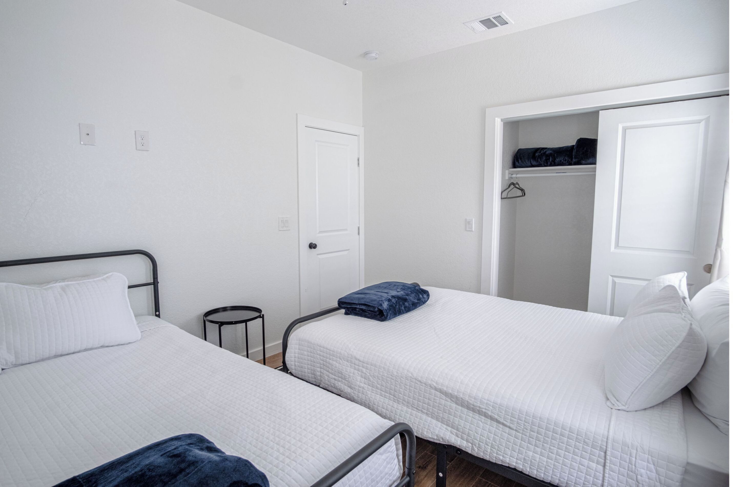A room with two single beds, white walls, a white door, and a closet with hanging clothes.