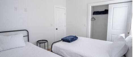 A room with two single beds, white walls, a white door, and a closet with hanging clothes.