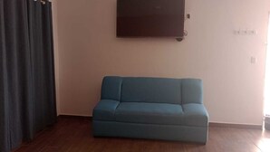 Quarto com sofá azul, cortina escura, TV fixada na parede e interruptor de luz.