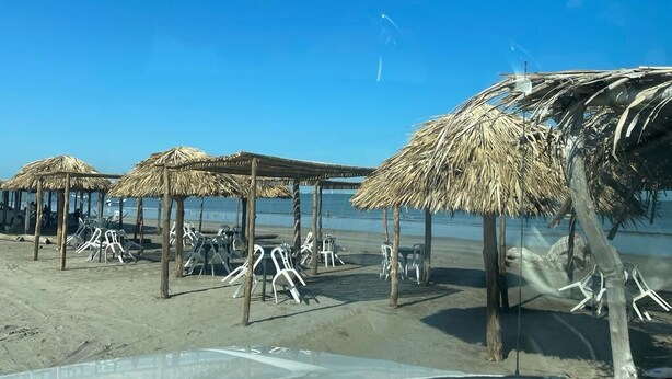 Uma praia com guarda-sóis de palha e cadeiras de plástico brancas.