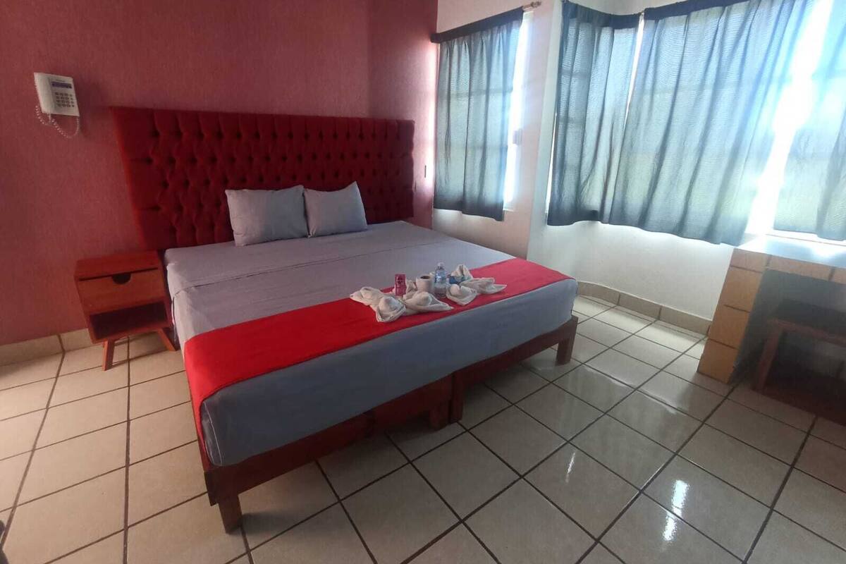 A hotel room with a large bed, a red headboard, a wooden nightstand, and a window with curtains.