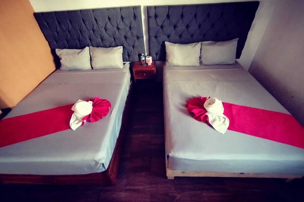 A hotel room with two beds, each with a red and white bedspread and a white pillow. There is a wooden nightstand between the beds with a bottle and a glass.
