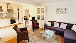 A modern living room with a sofa, two armchairs, a glass coffee table, and a kitchen area with white cabinets and a dining table. - Seesternchen Fewo Nr. 1 (St. Peter-Ording)