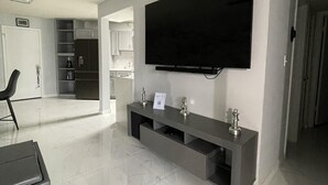 En moderne stue med et fladskærms-tv på væggen, et gråt konsolbord med pynteting, og et køkken i baggrunden.