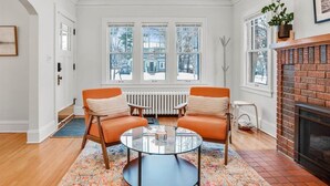 Ein Zimmer mit zwei orangefarbenen Sesseln, einem Glastisch, einem Kamin und Blick durch große Fenster ins Freie.